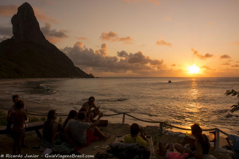 Imagem de turistas sentados e deitados curtindo o lindo por do sol em Noronha.